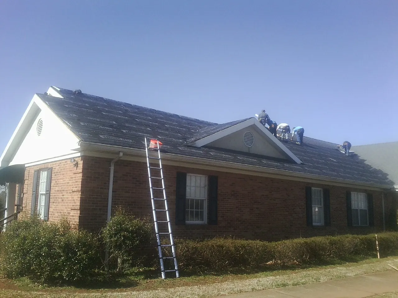 Crew tearing shingles off a brick ranch roof with a ladder set — Emergency Roof Repair work by our crew in Gilcrest