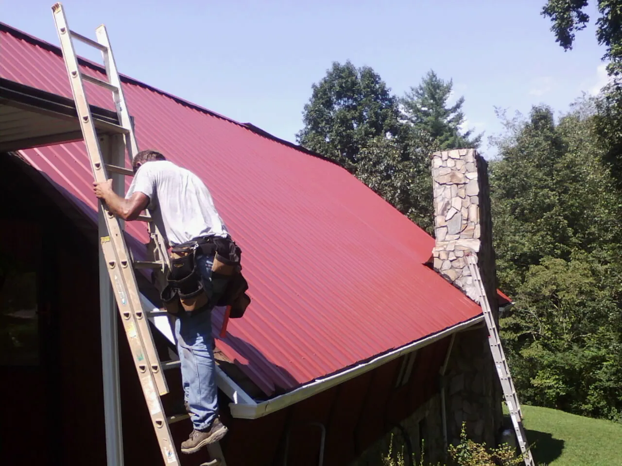 Roofer on a ladder beside a red metal roof and stone chimney — Soffit & Fascia Repair by Roofing Brothers in Gilcrest